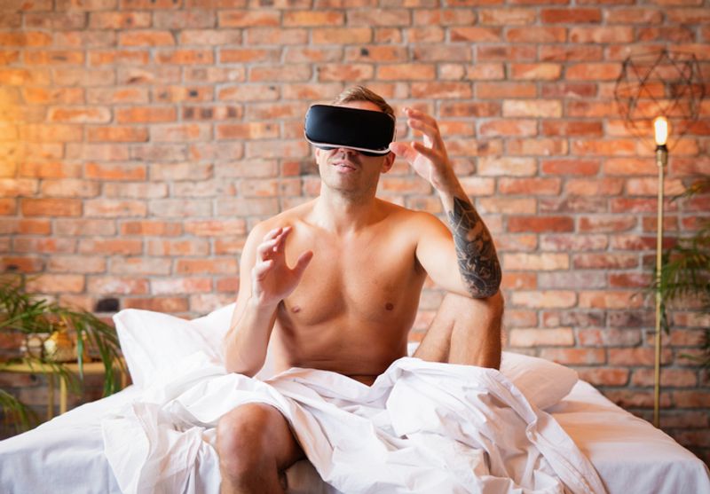 A shirtless man sits on the edge of bed covered in a sheet. He wears black virtual reality glasses and holds his hands in a questioning manner.