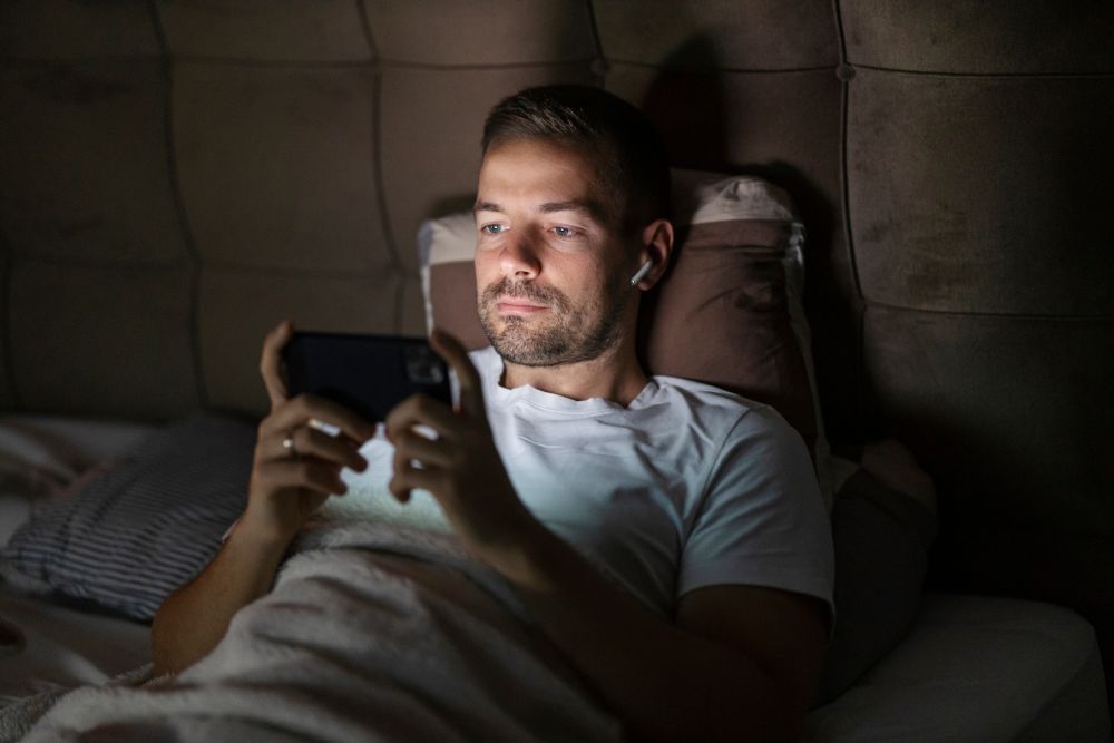 Man lying in bed with earbuds watching videos on smartphone