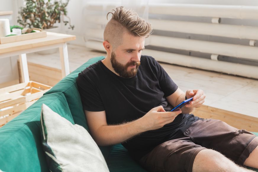 Man with faux-hawk sitting on green couch intently staring at smartphone