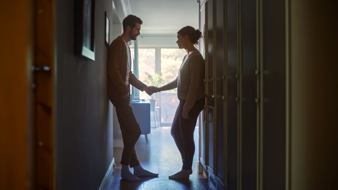 couple standing in backlit hallway across from each other while holding hands somberly